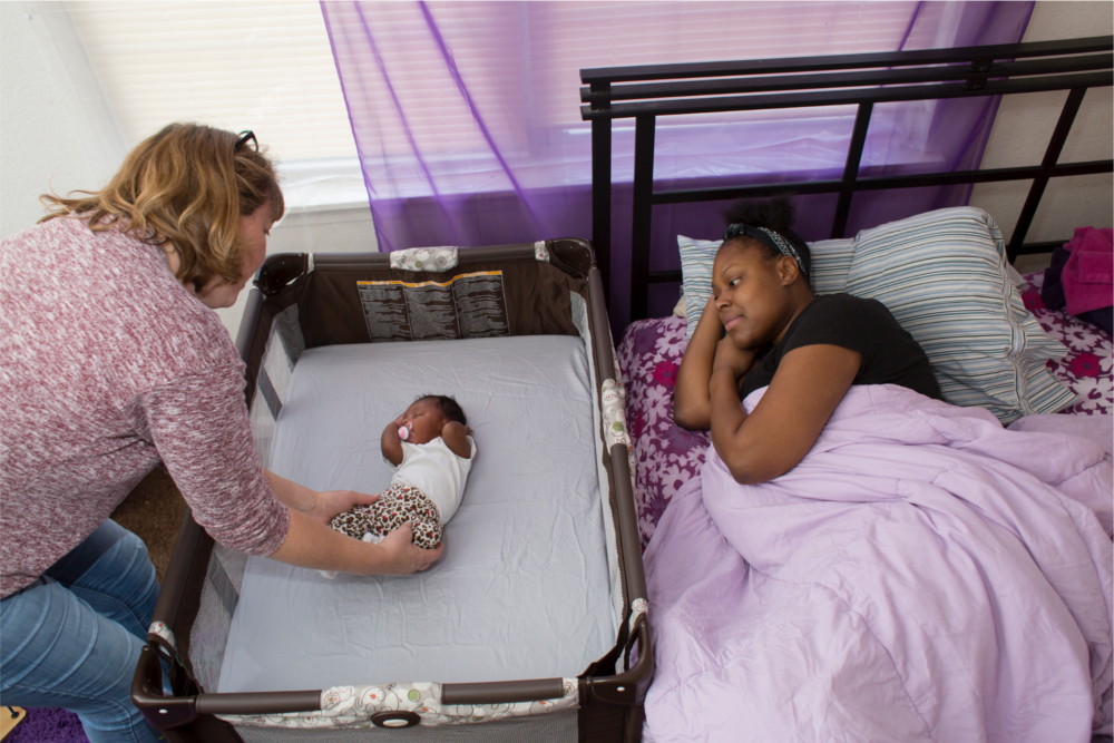 Woman laying in bed while her newborn baby is tended by a nurse in an adjoining crib.